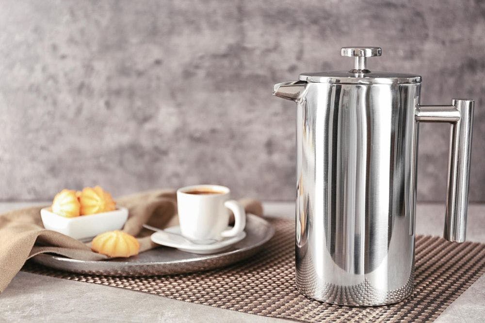 French press made up of stainless steel is placed near to the plate on which coffee is served